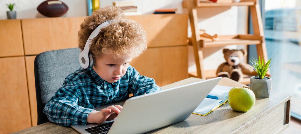 curly kid typing on laptop near apple while listening music in wireless headphones
